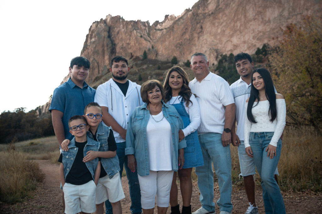 An outdoor family portrait features eight people posing and smiling on a dirt path with a mountain in the background.