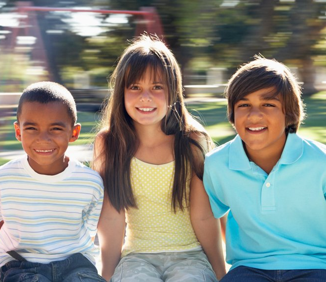Three children sitting outdoors, smiling at the camera on a sunny day, with a blurred playground in the background.