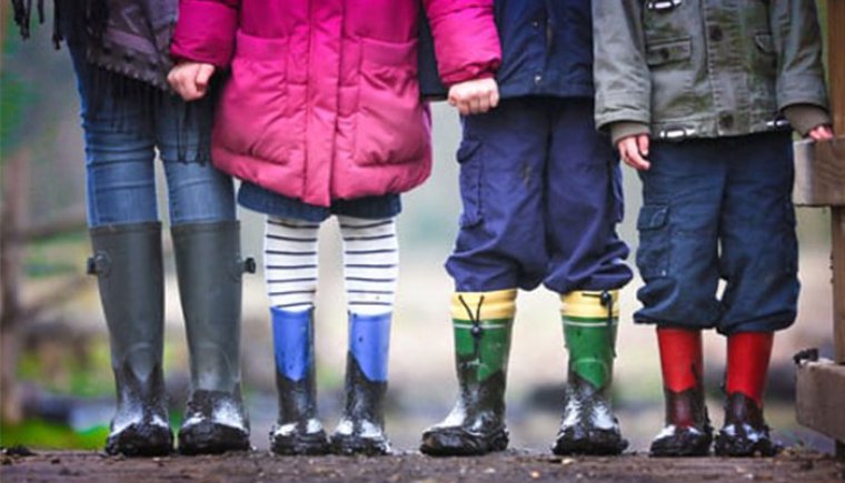 Four kids in rain boots stand together outside.